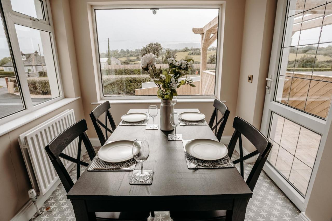 Dining table set for four with flowers, near large window overlooking countryside.