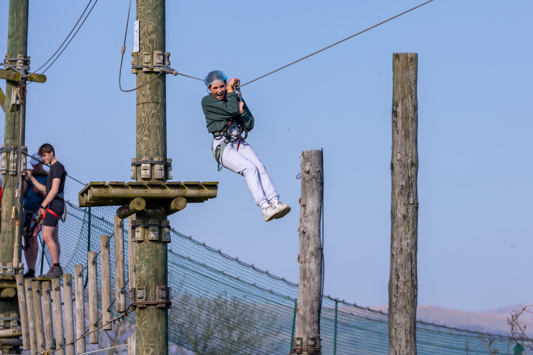 Person zip-lining with safety gear, smiling, against a clear blue sky.
