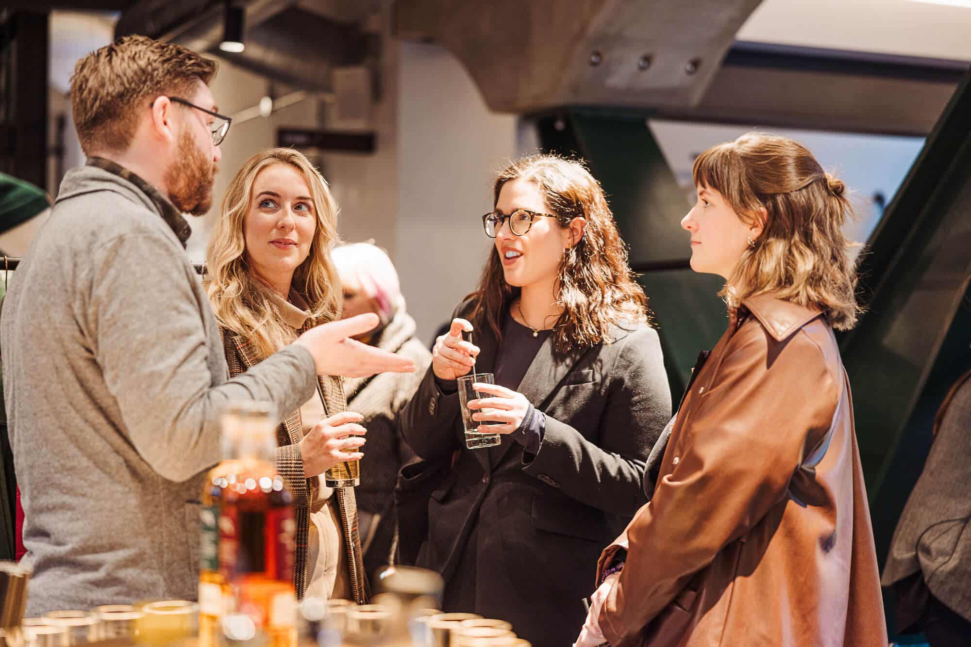 Four people conversing indoors, holding drinks, with bottles on a table.
