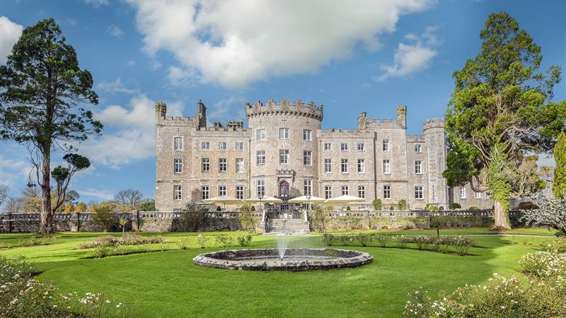 A large stone castle with a manicured lawn and a circular fountain in the foreground.