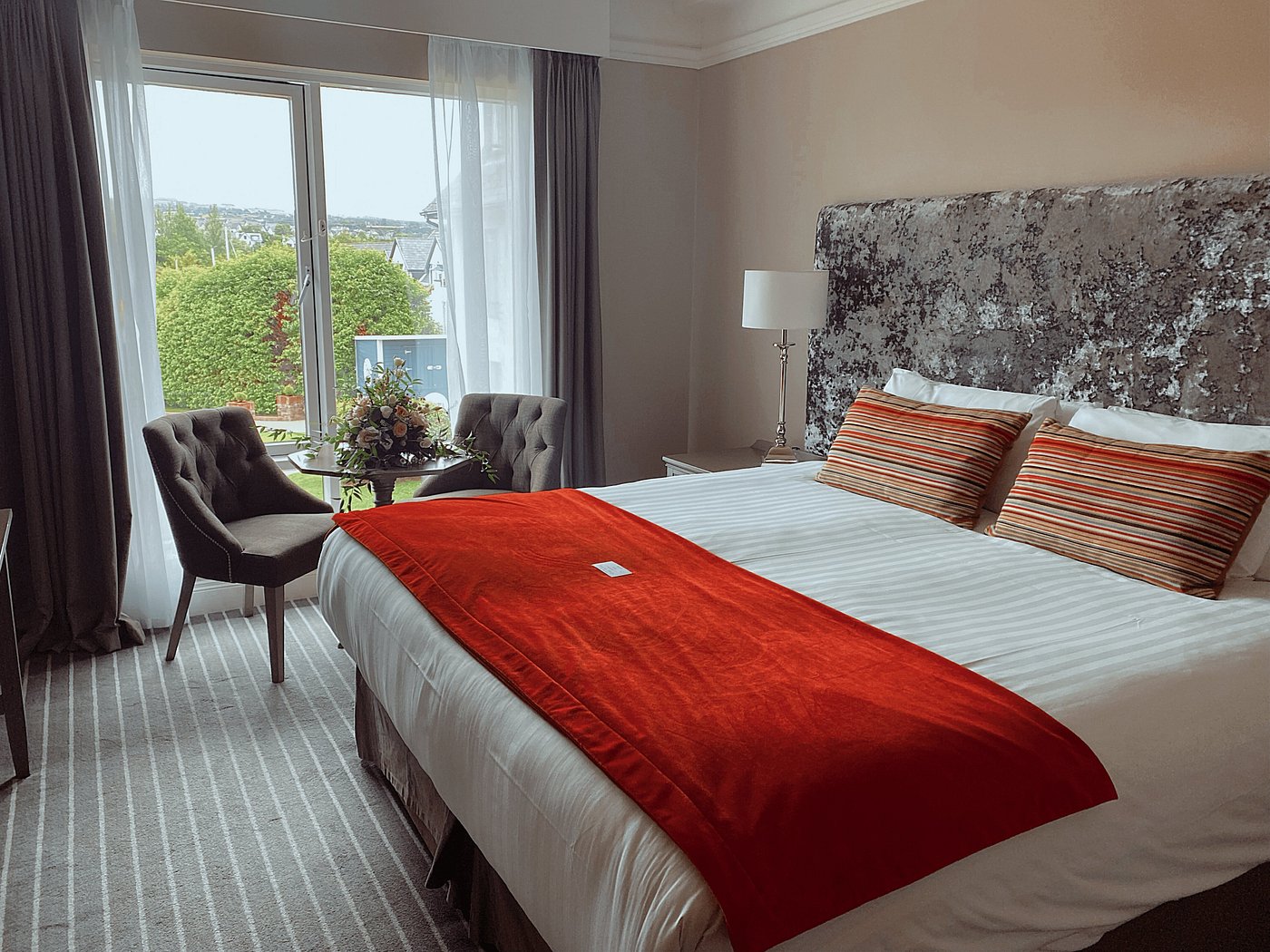 Hotel room with a large bed, red throw, and window view of greenery.