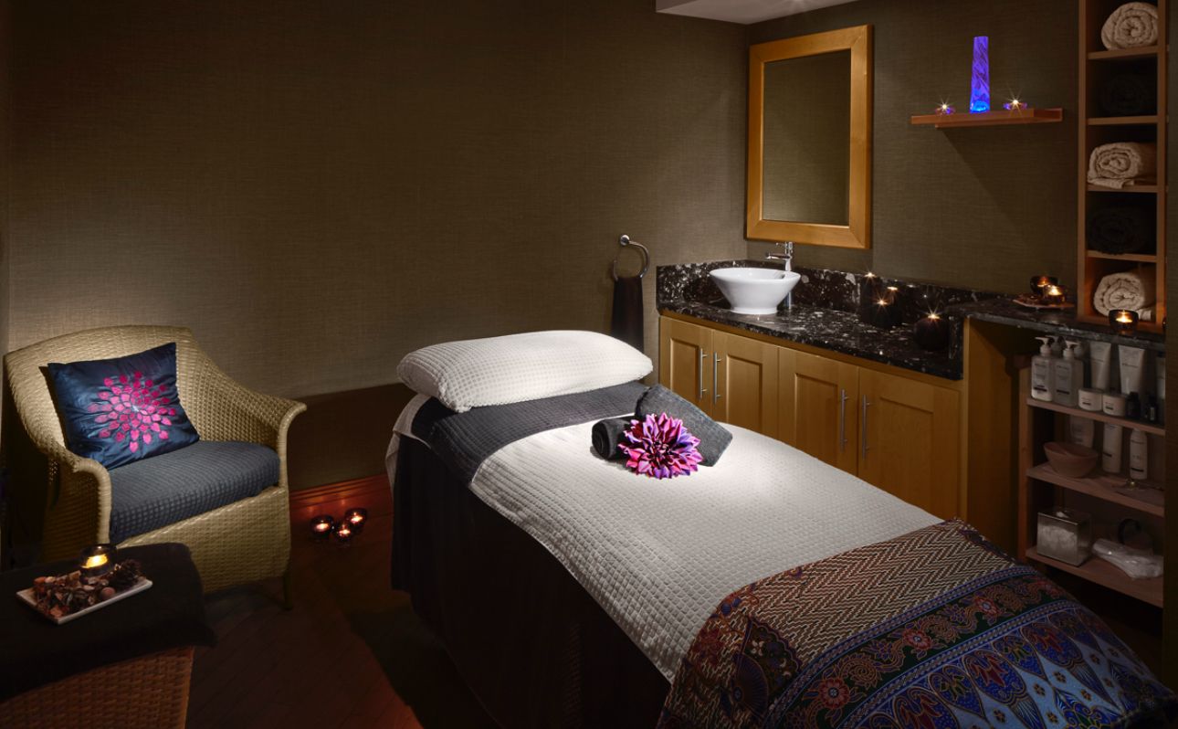 A cozy spa room with a massage table, candles, and a wicker chair.