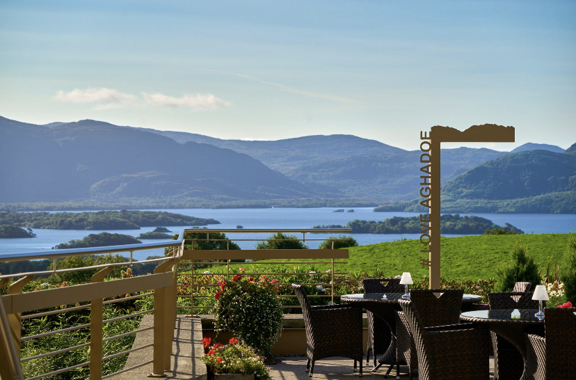 A scenic view of a lake and mountains from an outdoor patio with tables and chairs.