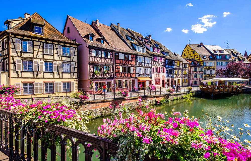 Vibrant flowers on a bridge overlook a canal lined with colorful, half-timbered houses under a clear blue sky.