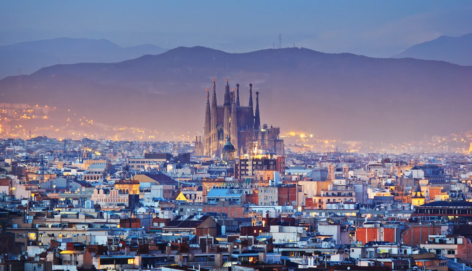 The Sagrada Familia church rising above the cityscape of Barcelona at dusk, with mountains in the background.