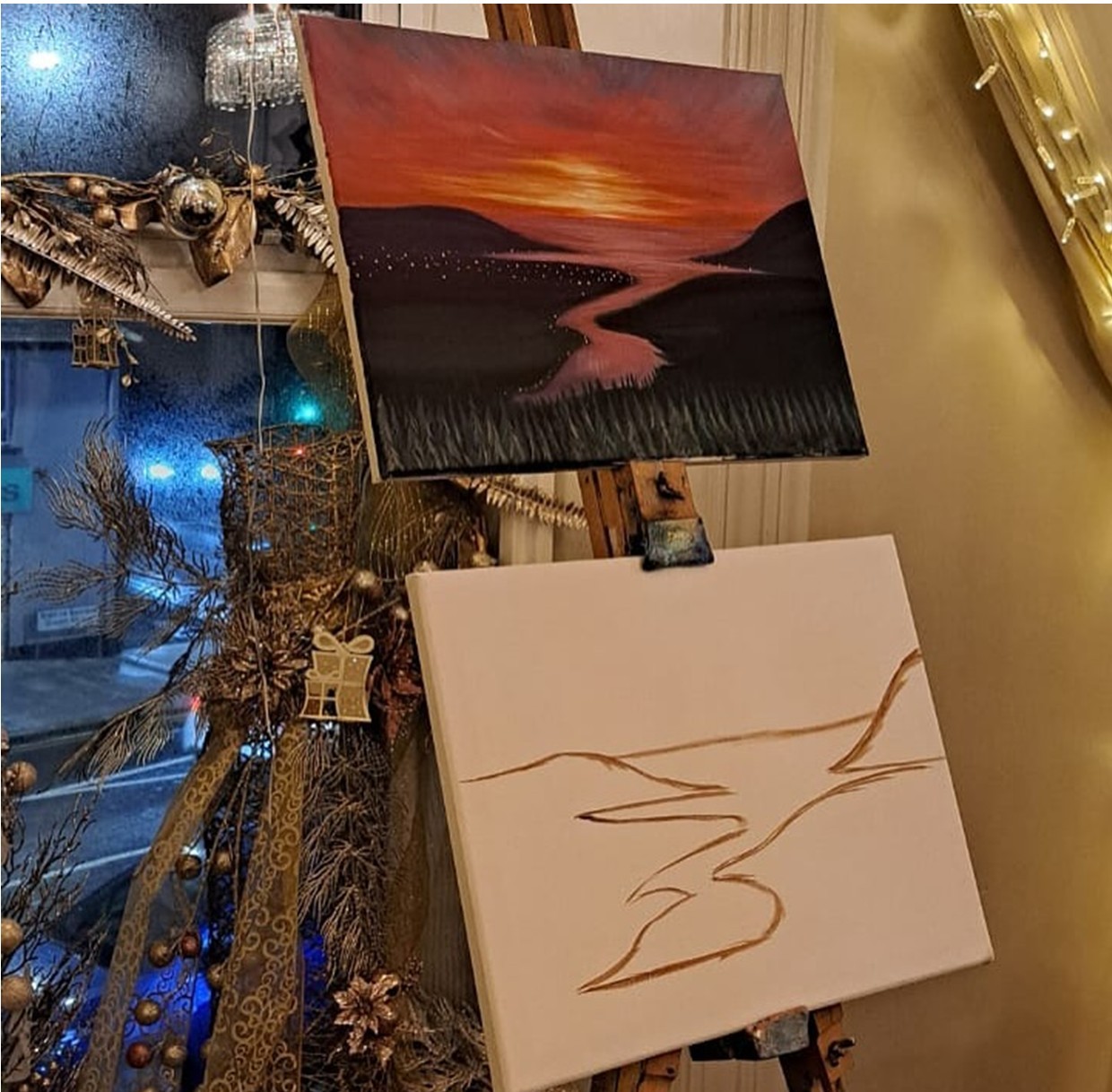 Two paintings on an easel, one finished and one sketch, near a decorated window.
