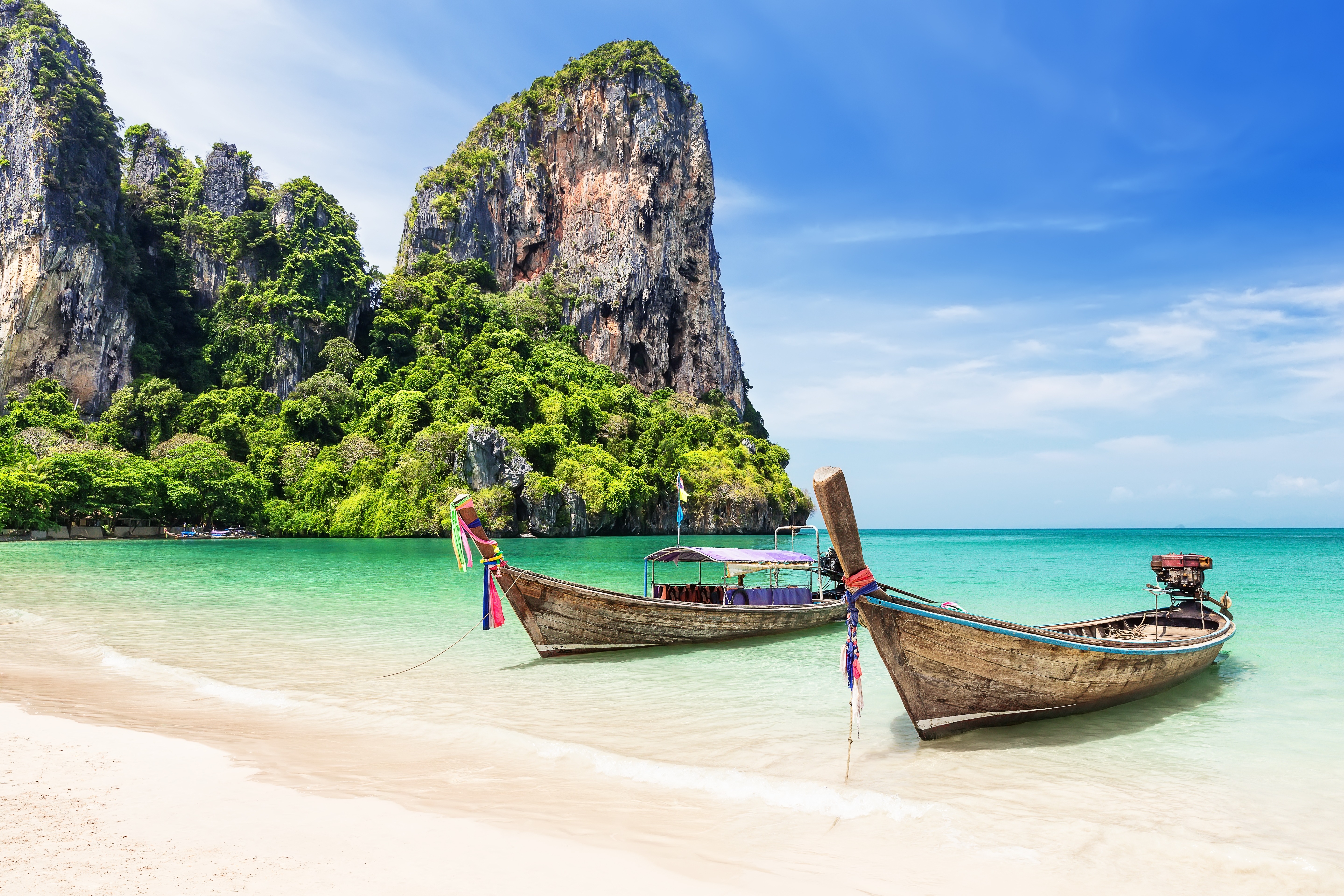 Two wooden long-tail boats rest on a sandy beach next to a large, jungle-covered limestone cliff.