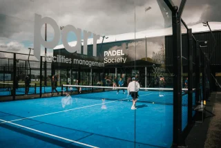 People play padel on a bright blue court, viewed from behind a glass door with logos.