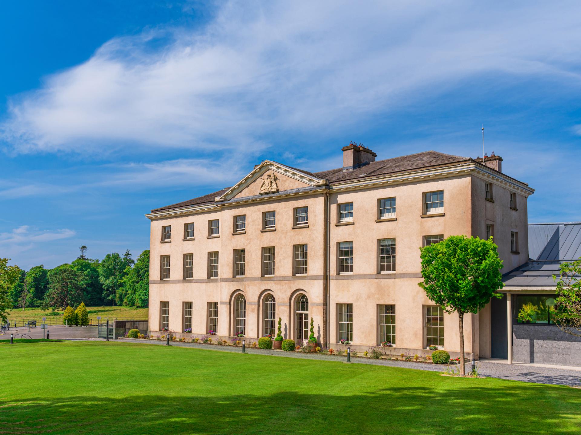 Large beige building with arched windows, surrounded by green lawn and trees under a blue sky.