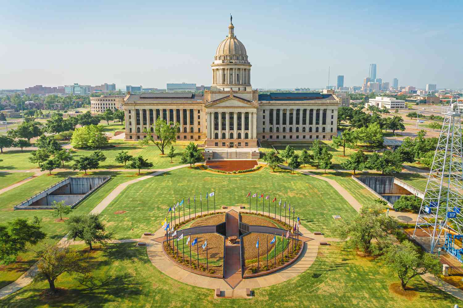 Large building with dome, surrounded by flags and greenery, city skyline in background.