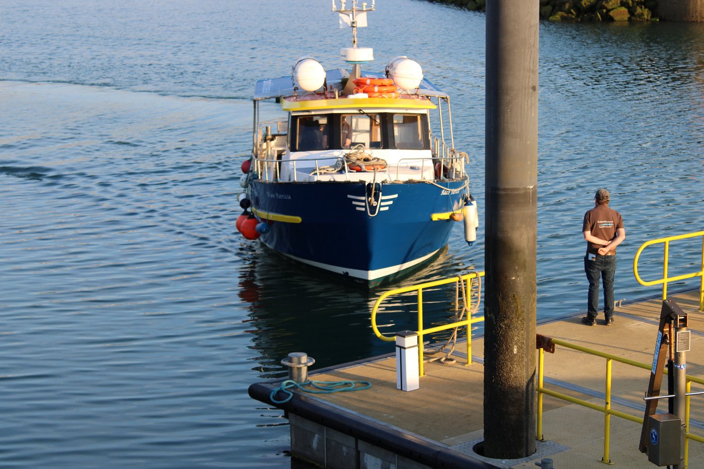 A blue boat approaching a dock with a person standing nearby.