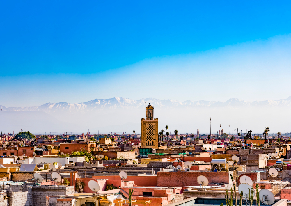 A cityscape with a prominent tower, with snow-capped mountains in the distance under a clear blue sky.