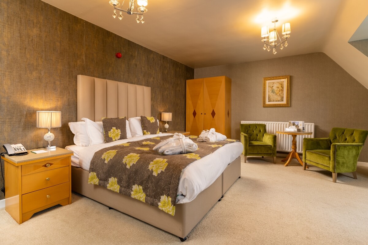 A spacious hotel room with a large bed, wooden wardrobe, and two green velvet armchairs.