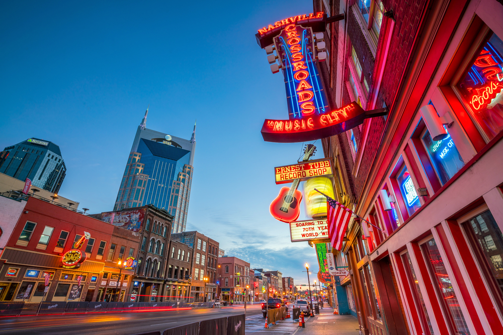A downtown Nashville street at dusk, with glowing neon signs, historic buildings, and modern skyscrapers.