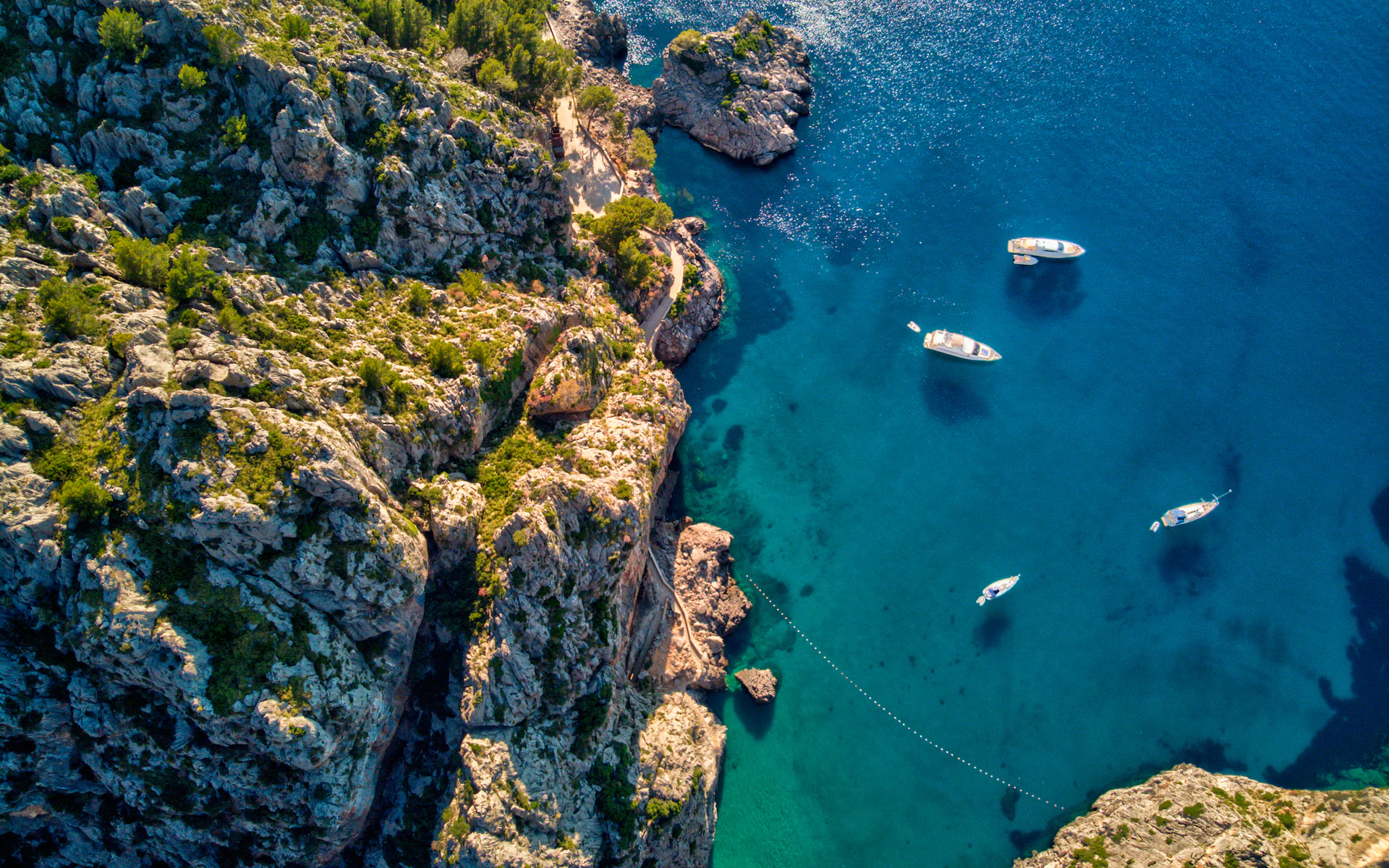 Aerial view of rocky coastline with boats on clear blue water.