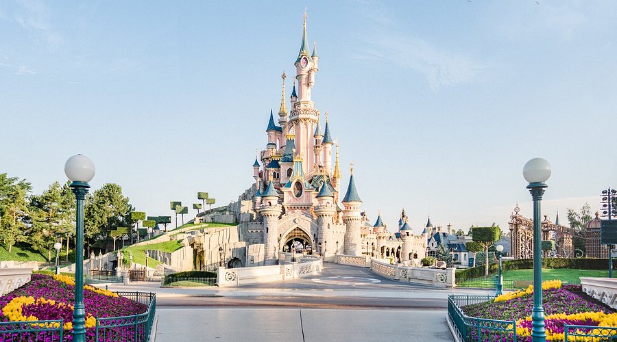 Colorful castle with spires, surrounded by gardens and pathways under a clear sky.