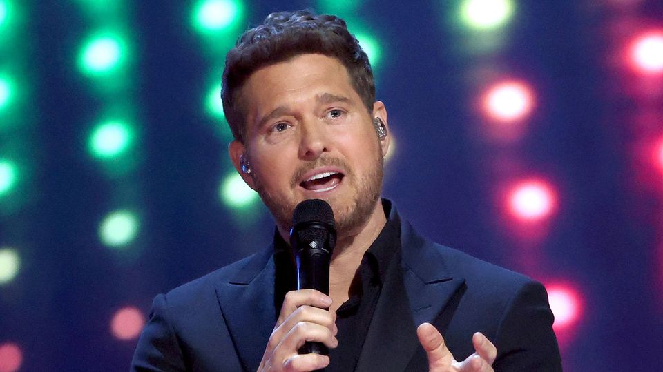 A man in a dark suit singing into a microphone with a background of blurred colored lights.