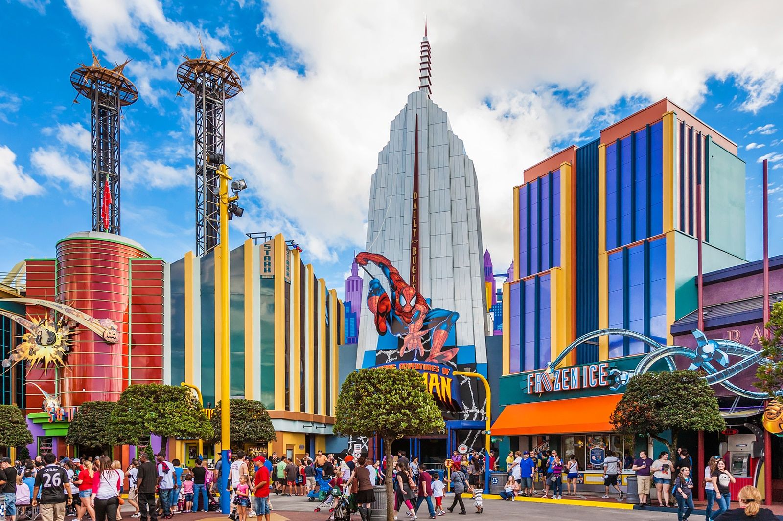 A bustling street at a theme park with colorful, comic-book style buildings and a large Spider-Man figure.