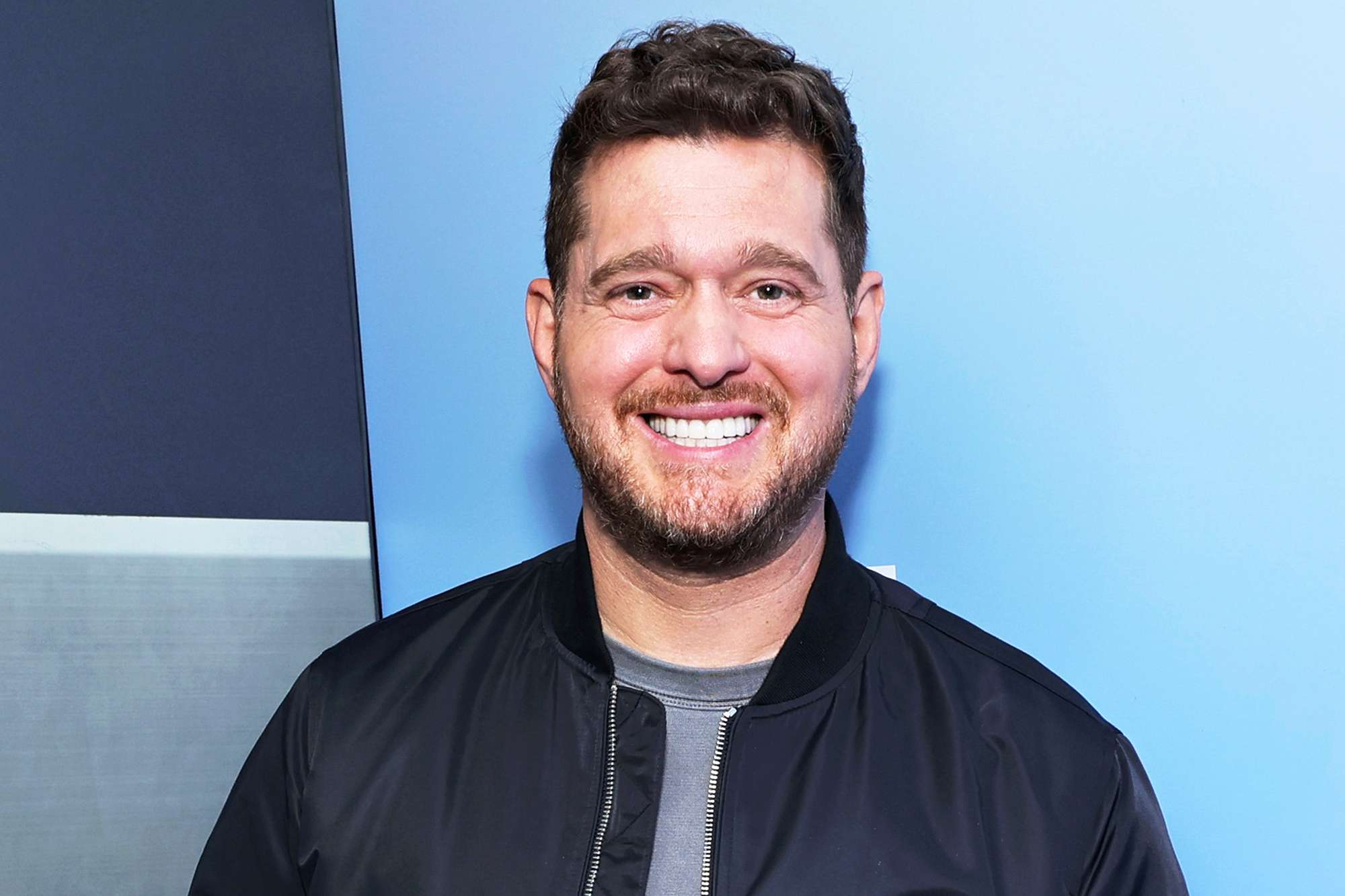 A headshot of singer Michael Bublé smiling while wearing a black jacket against a blue background.