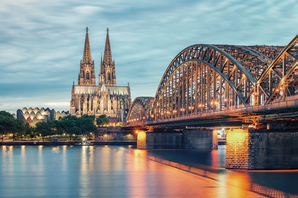 The Hohenzollern Bridge and Cologne Cathedral over the Rhine river at sunset.
