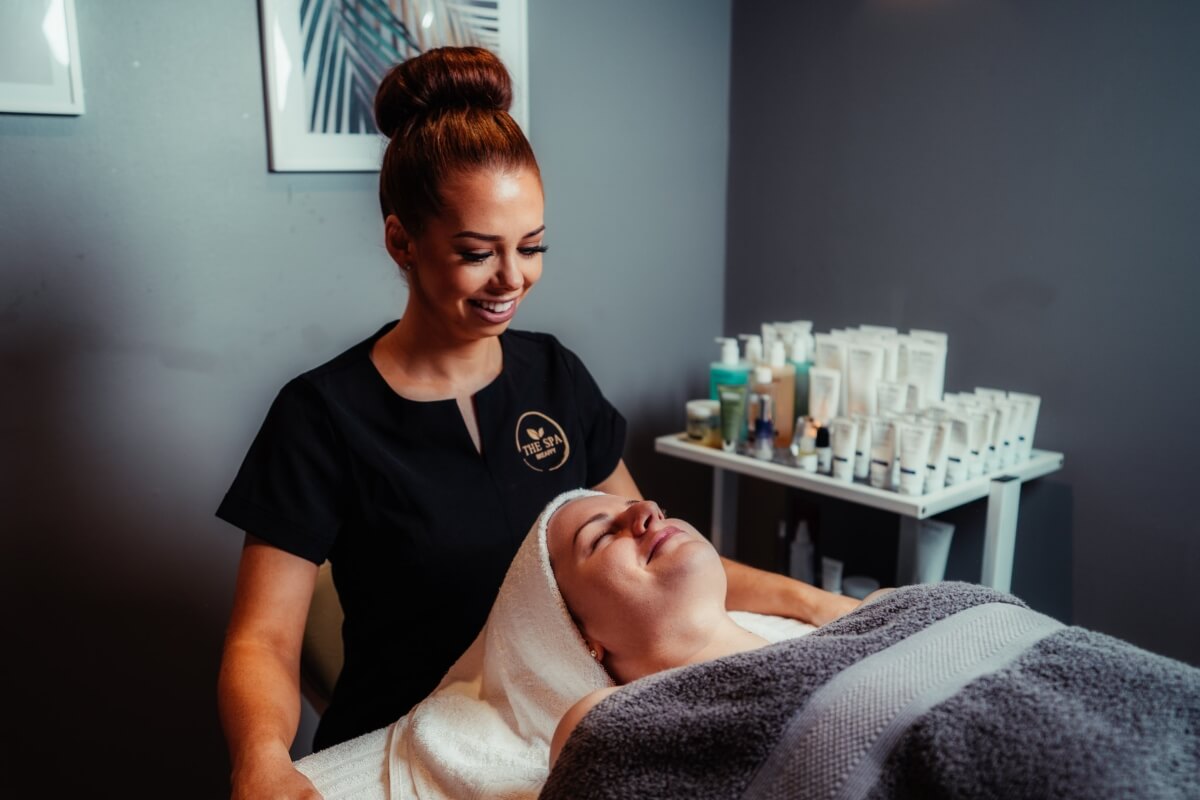 A smiling esthetician gives a facial to a relaxed client lying on a treatment bed in a spa.