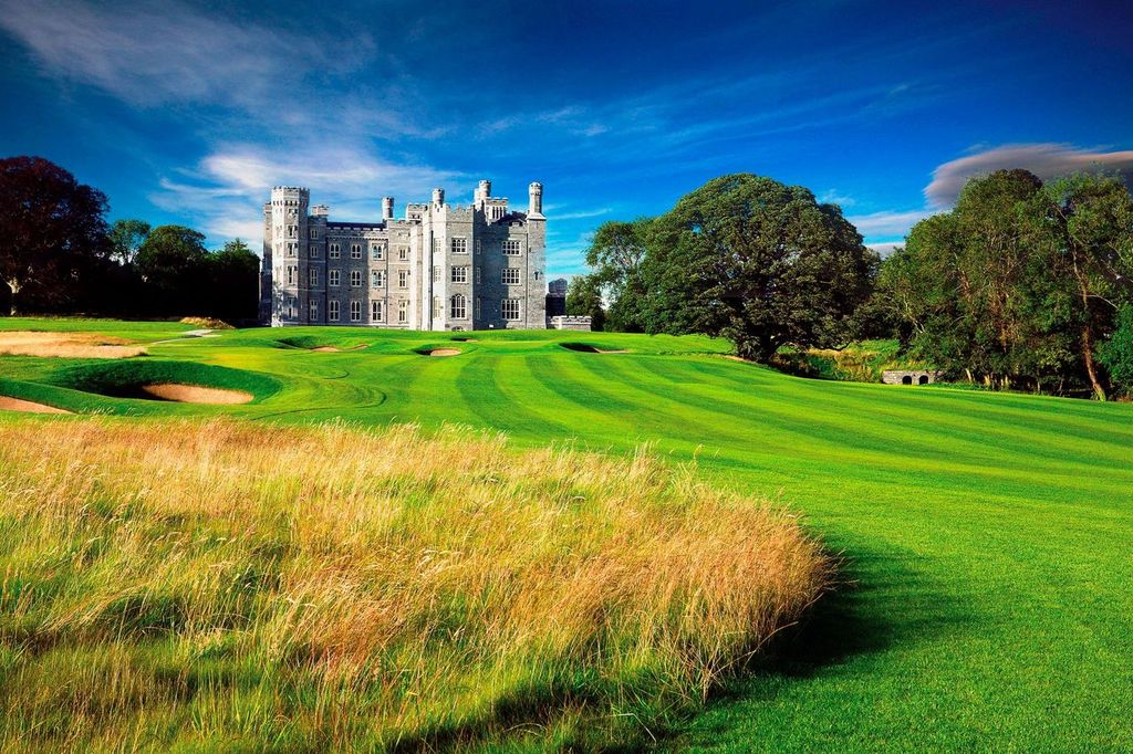 A large stone castle sits behind a lush green golf course under a vibrant blue sky.
