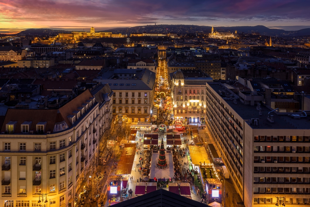 An aerial view of a bustling Christmas market lining a city street at dusk, with the cityscape behind.