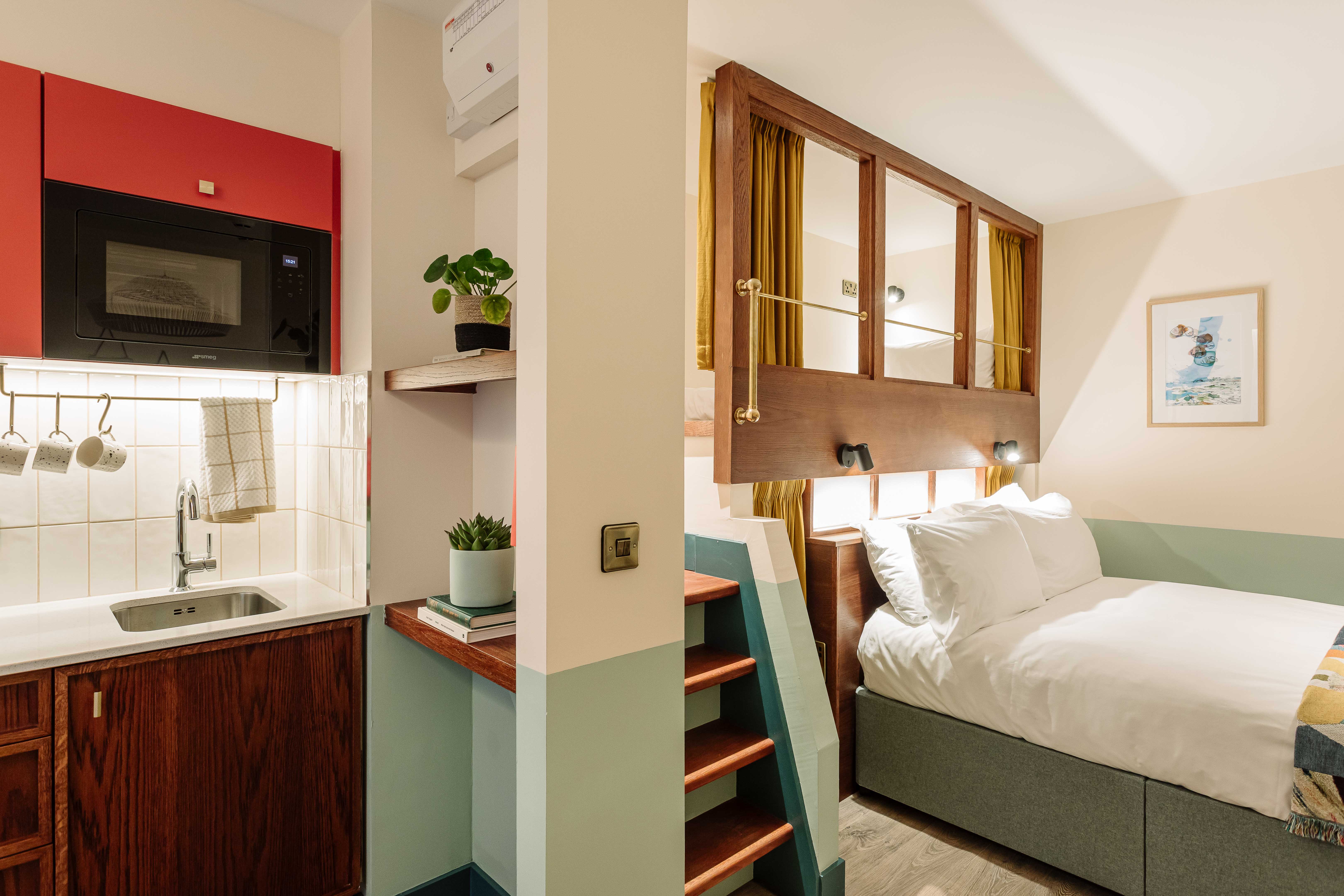 A compact, modern, split-level room showing a kitchenette on the left and a bed on the right.