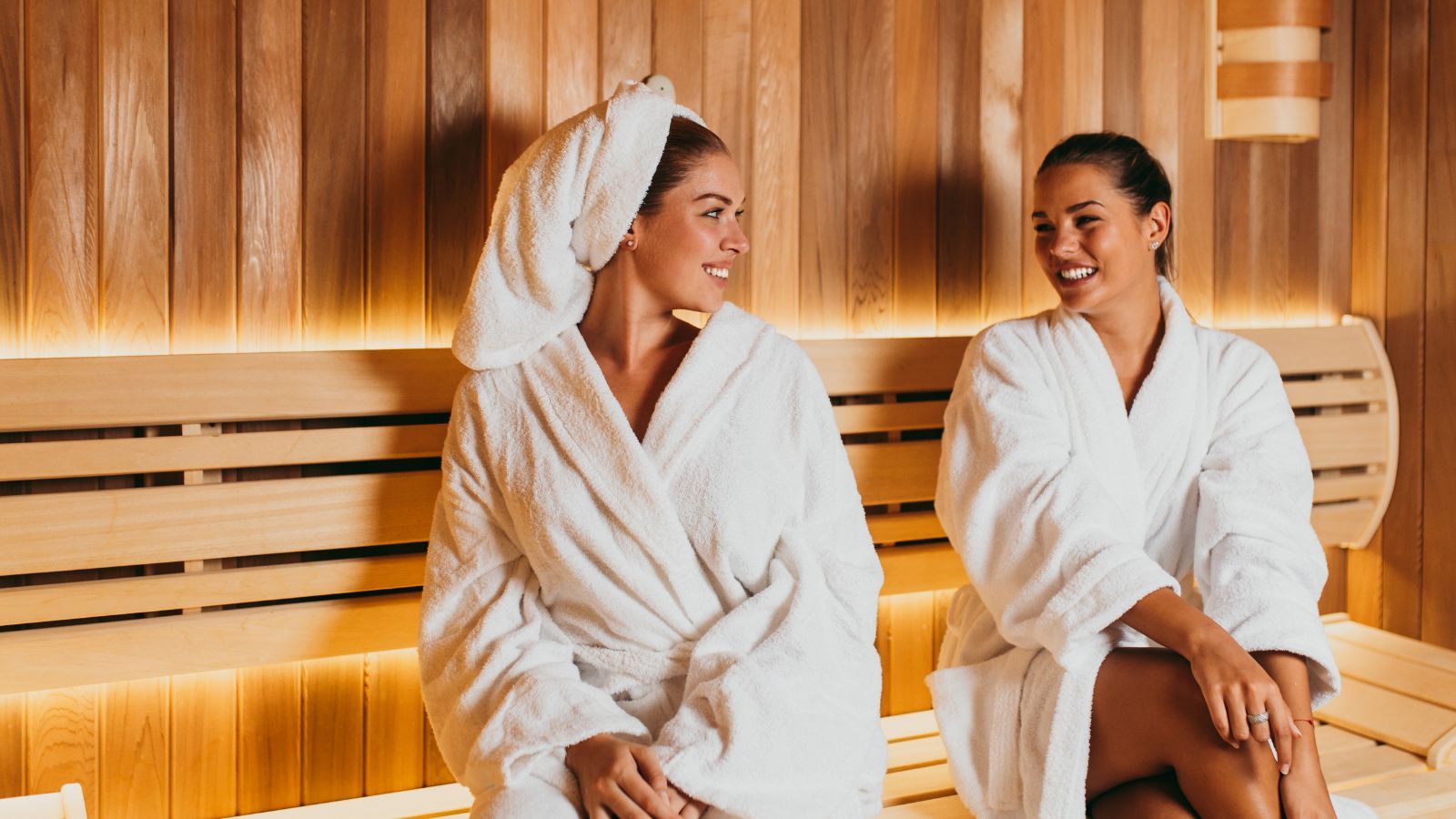 Two women in white robes sitting in a wooden sauna, smiling at each other.