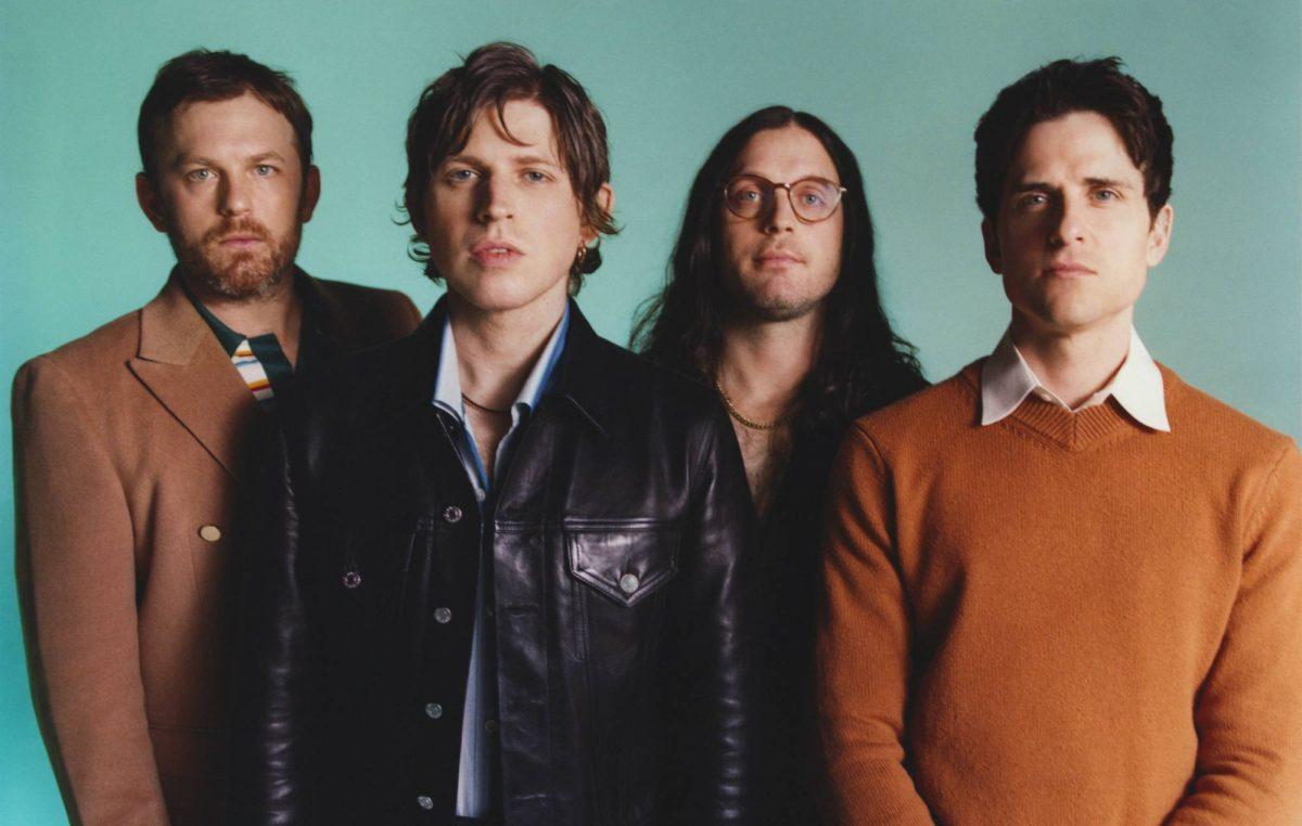 A group portrait of four men standing together against a plain teal background, looking at the camera.