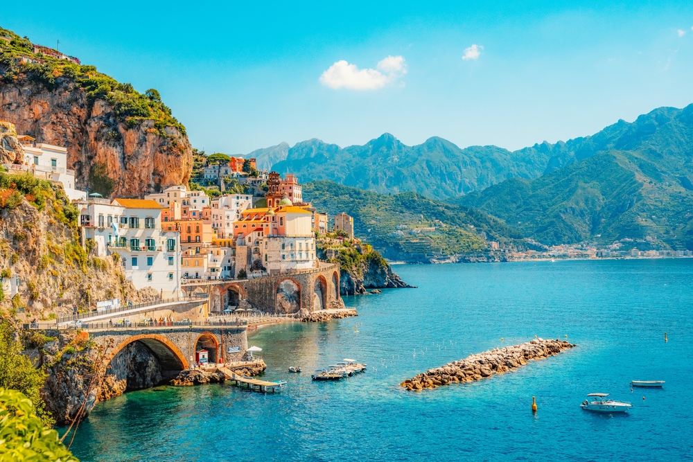 A colorful town built on a cliffside overlooking a bright blue ocean, with mountains in the background.