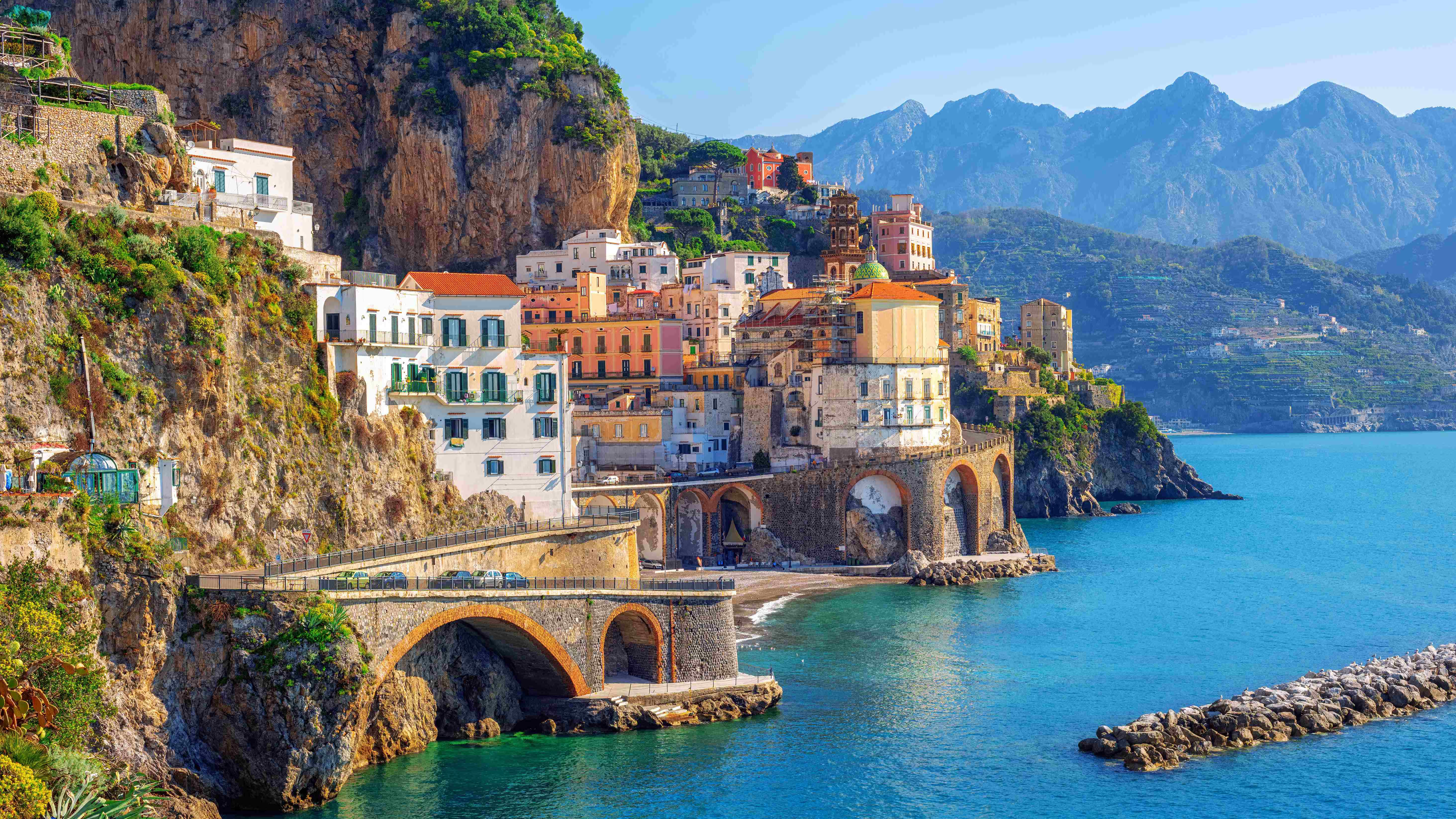 A colorful town built into a seaside cliff, with an arched bridge and mountains in the background.