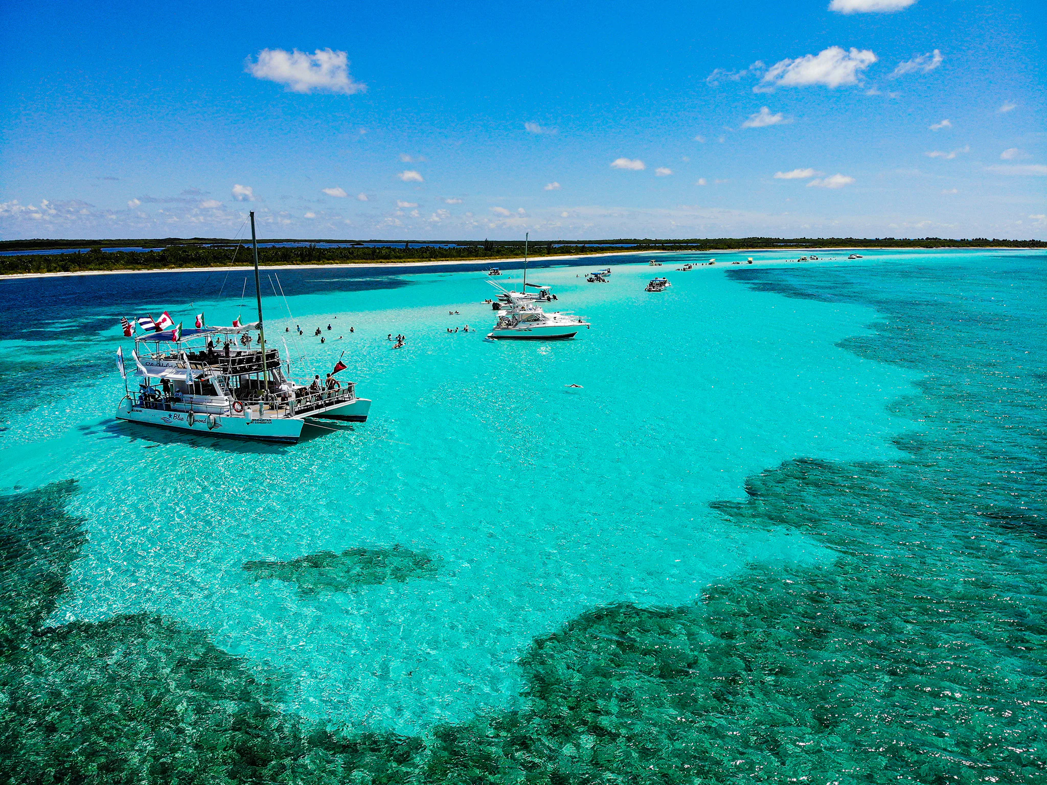 An aerial view of boats and people in shallow, turquoise tropical waters off a sandy coast.