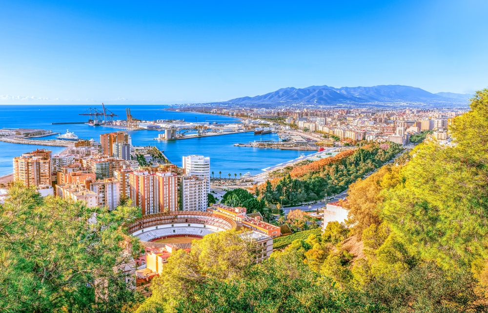 A high-angle, sunny day view of a coastal city with a large port, a bullring, and mountains.