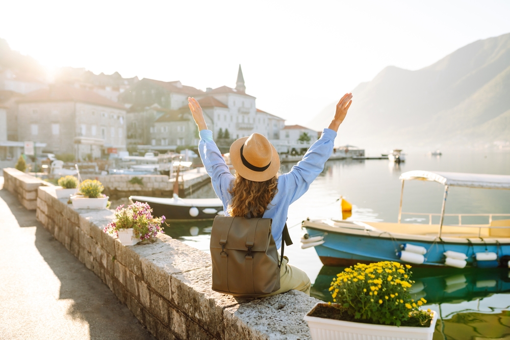 A woman with a backpack and hat sits on a stone wall, arms raised, overlooking a coastal town.