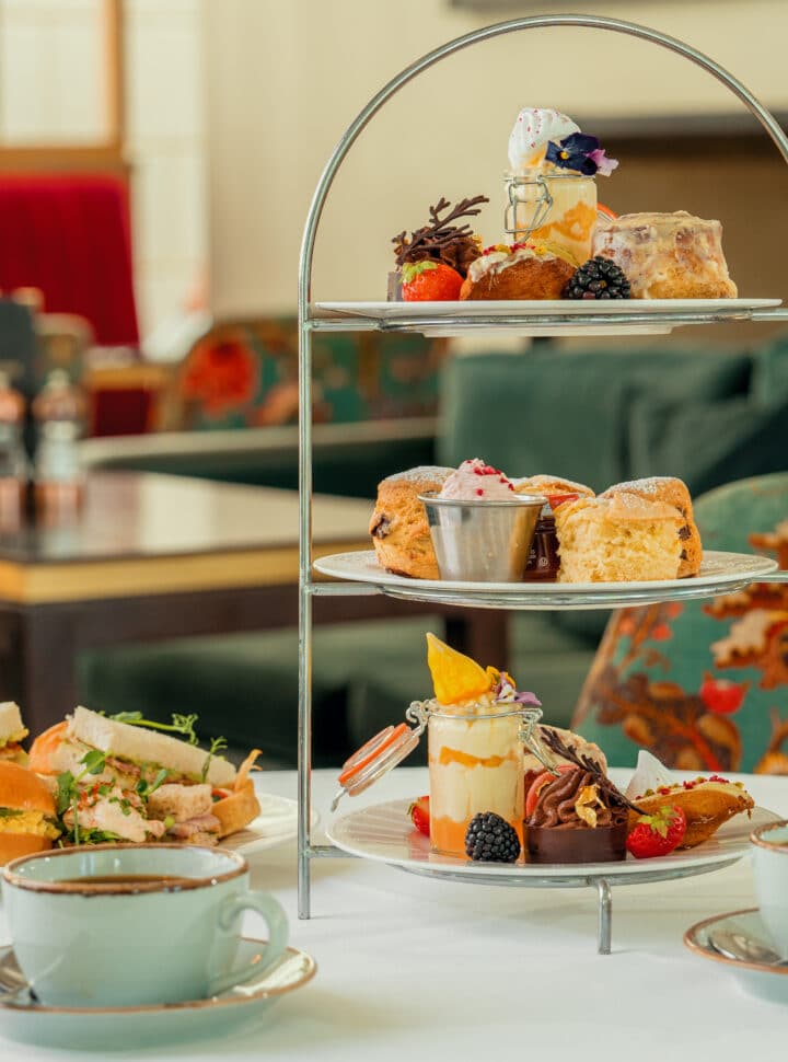 Three-tiered afternoon tea stand with pastries, sandwiches, and a cup of coffee on a table.