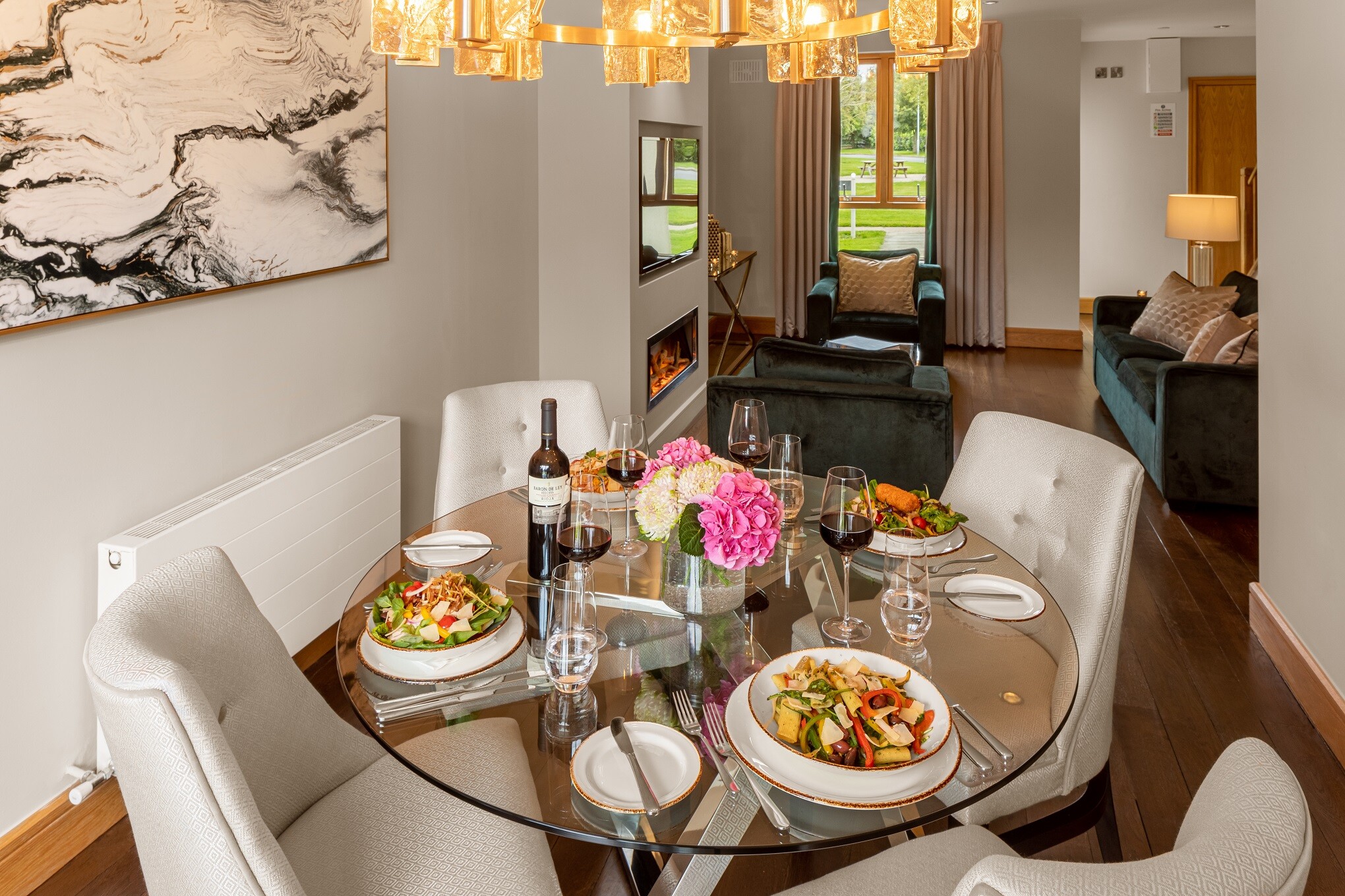 A modern dining room with a glass table set for a meal, overlooking a living area and fireplace.