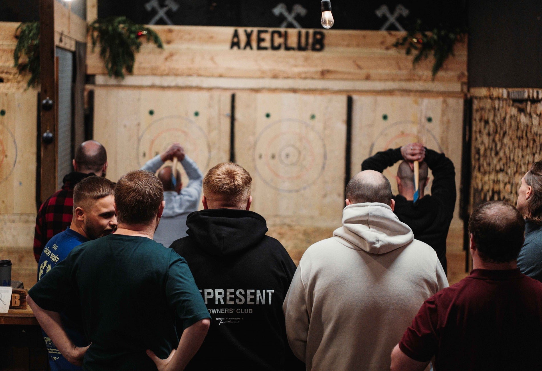 A group of men from behind watch others prepare to throw axes at wooden targets at an axe club.
