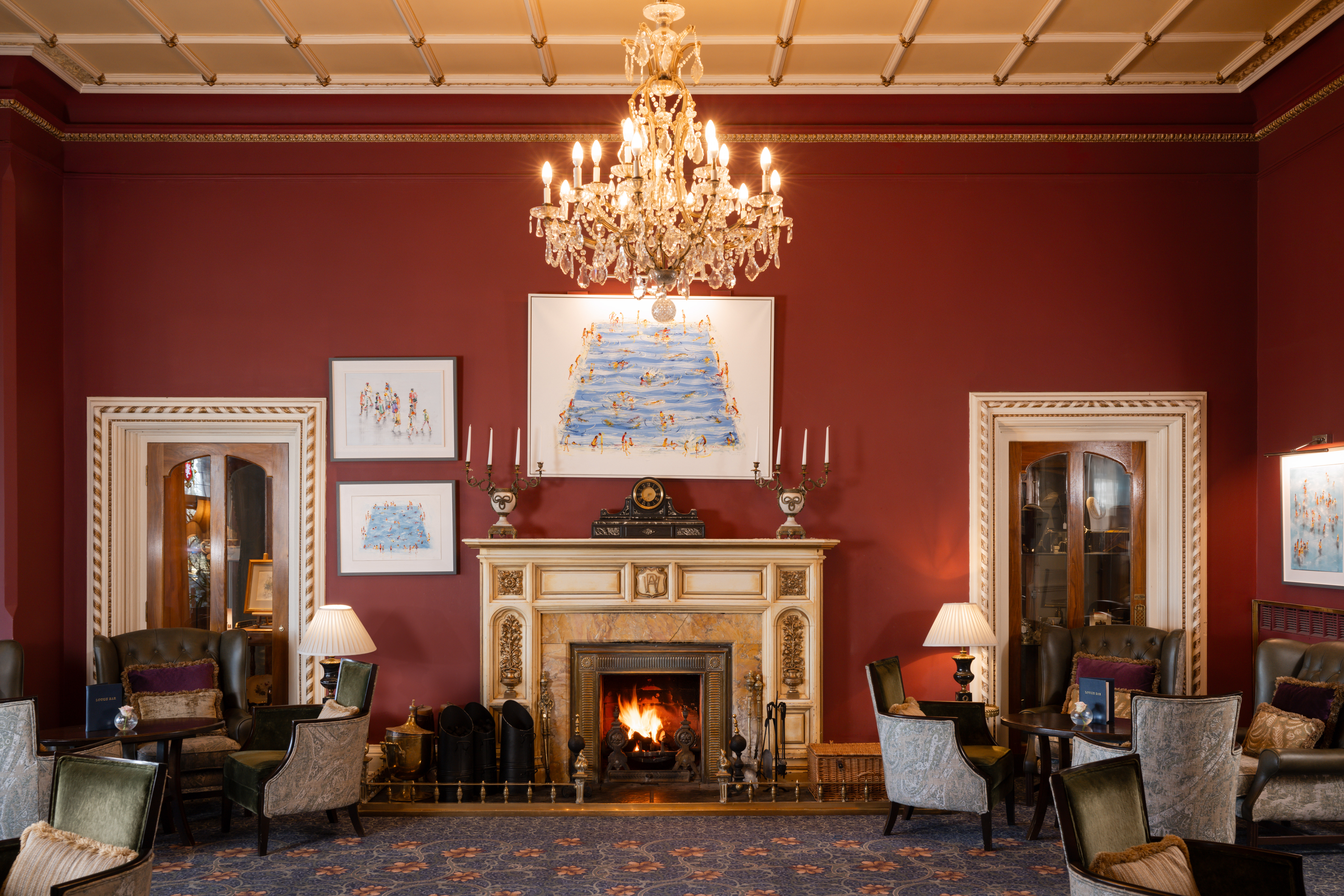 Elegant room with a chandelier, fireplace, armchairs, and artwork on burgundy walls.