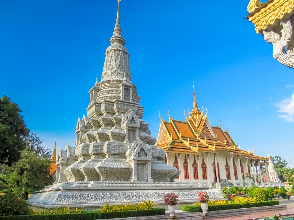 An ornate, white stupa and a temple with a golden roof sit in a garden under a blue sky.