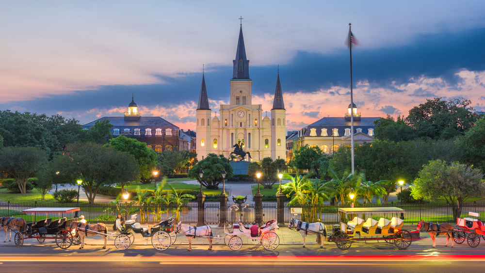 Horse-drawn carriages line a street in front of St. Louis Cathedral in New Orleans at dusk.