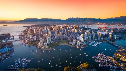 An aerial view of a downtown peninsula surrounded by water, with boats in the harbor and mountains beyond at sunset.