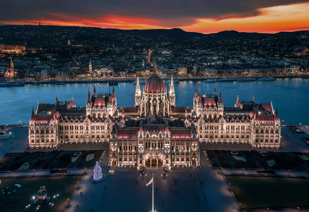 An aerial view of the illuminated Hungarian Parliament Building at dusk, with the city and a river behind.