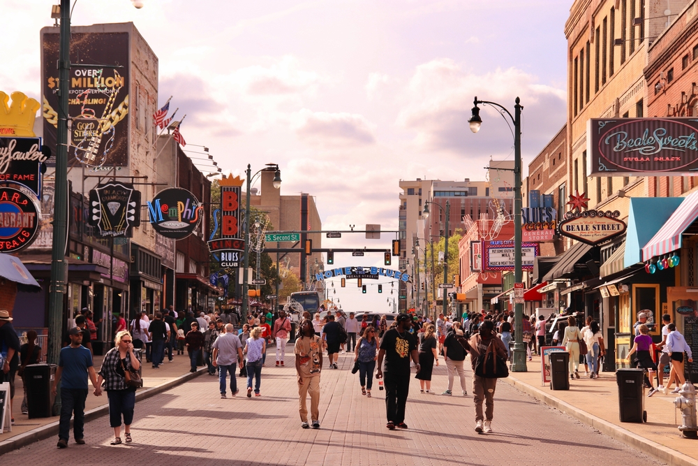 A crowd of people walks down a sunlit, brick-paved city street lined with buildings and neon signs.