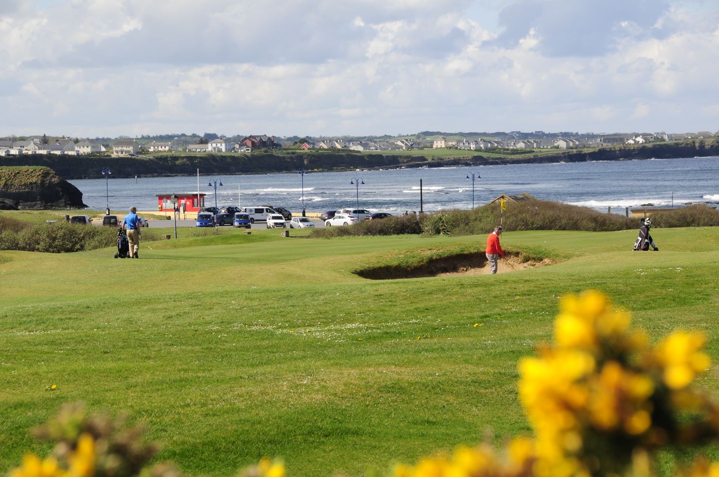 Golfers on a green coastal course, with the ocean and a town in the background.