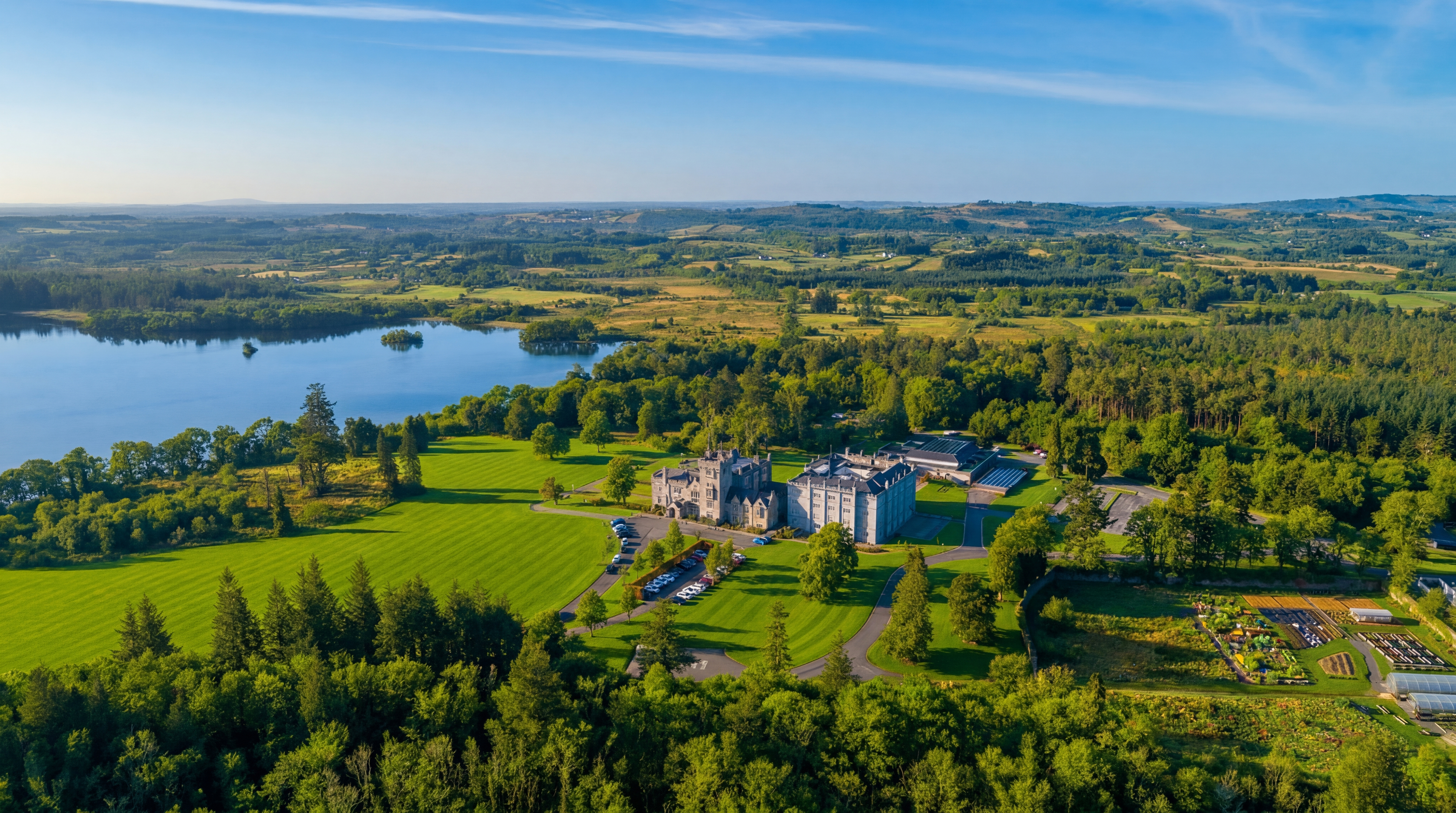 Aerial view of a large castle estate beside a lake, surrounded by lush green forests and rolling hills.