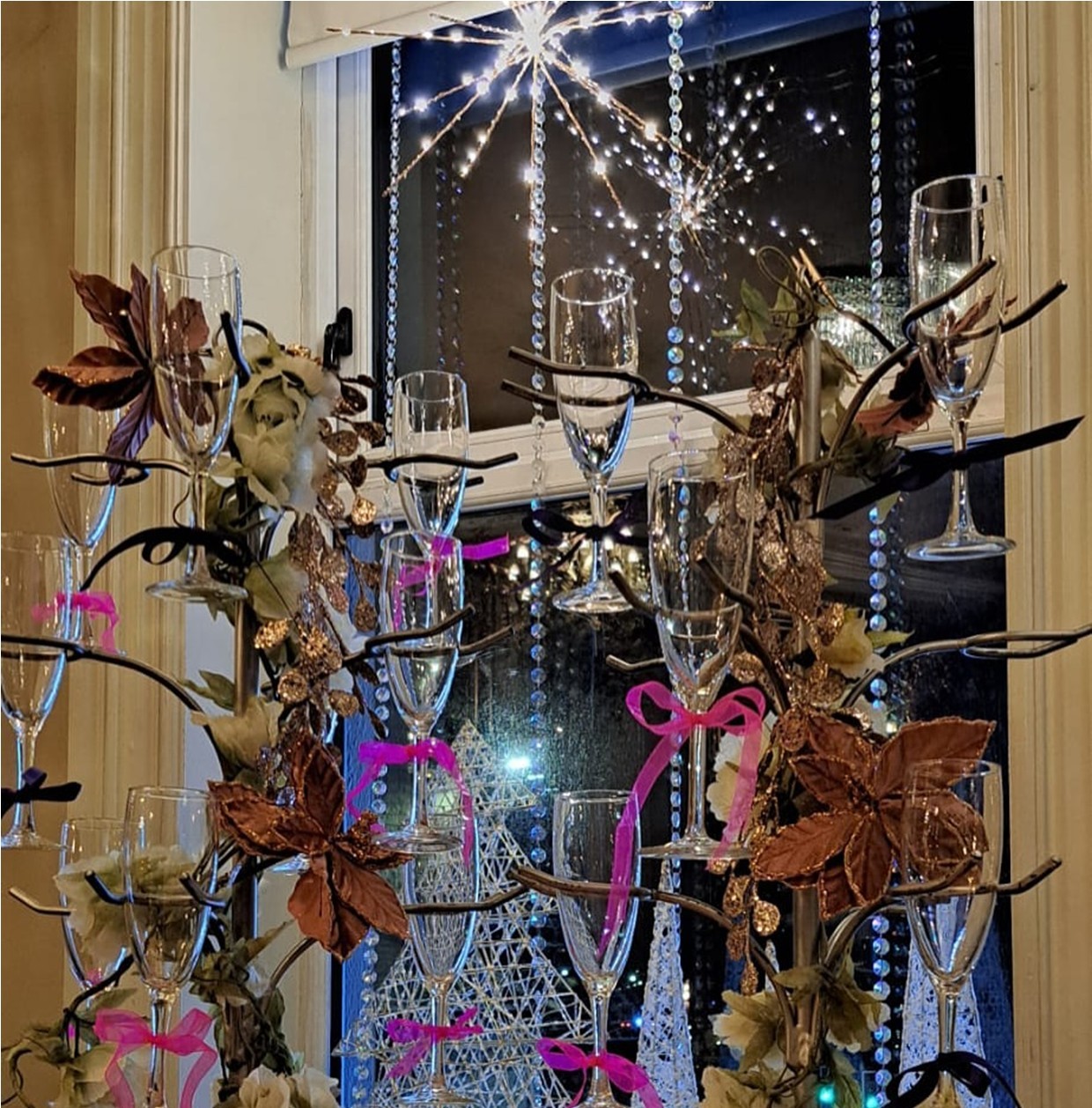 Decorative champagne glass display with flowers and ribbons, in front of a window with lights.