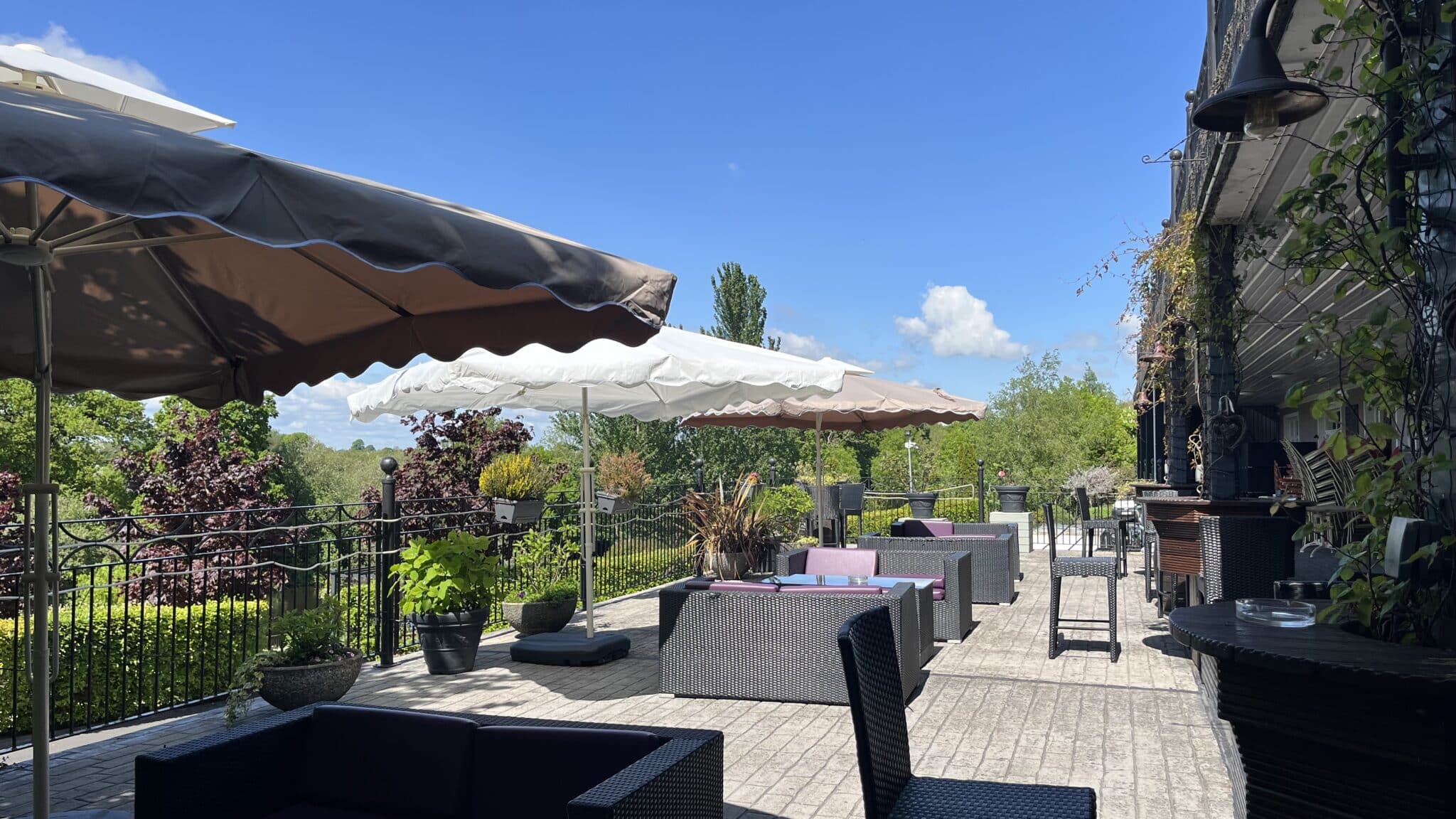 Outdoor patio with umbrellas, wicker furniture, and greenery under a clear blue sky.
