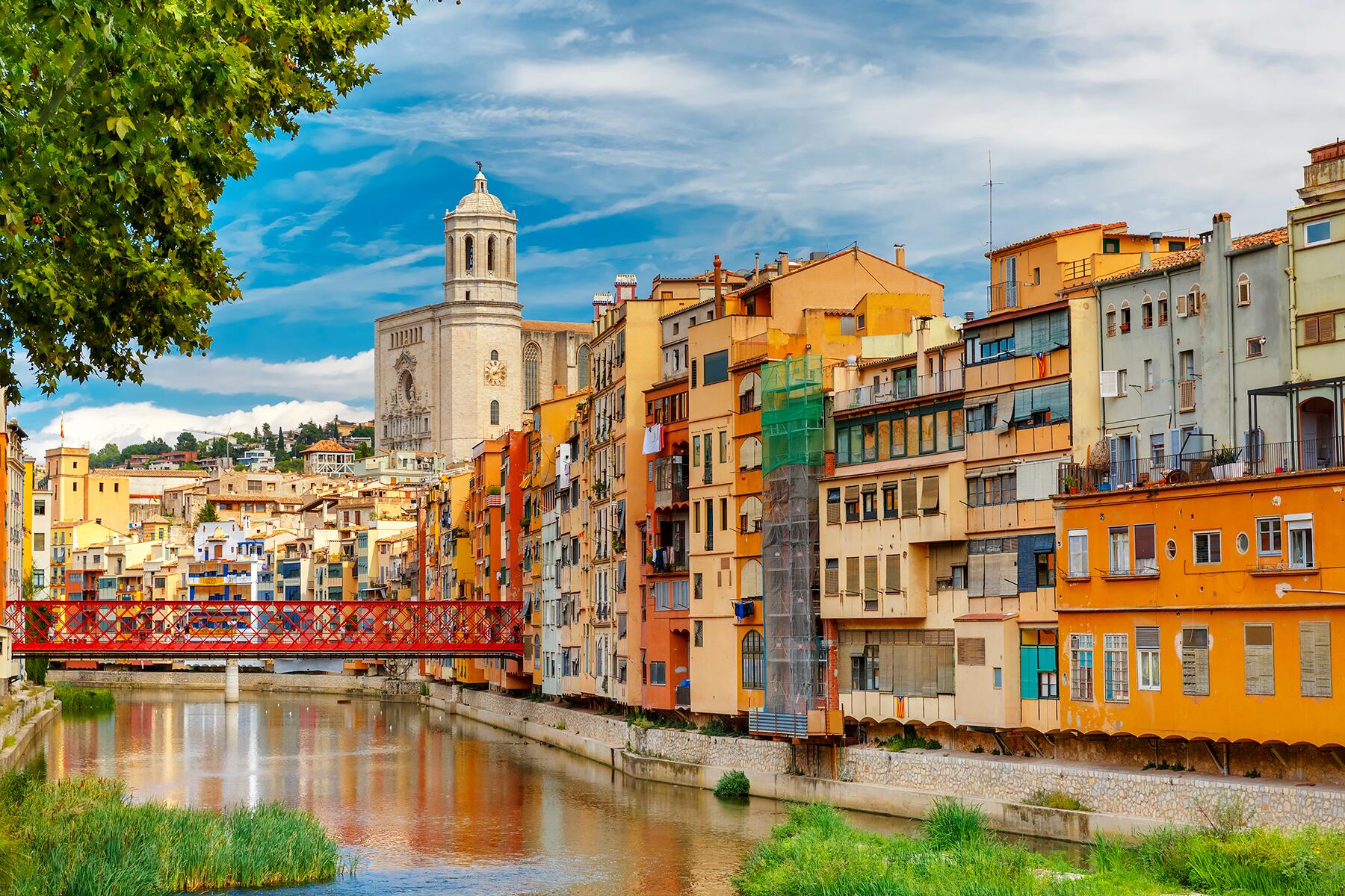 Colorful buildings along a river with a red bridge and a large cathedral in the background.