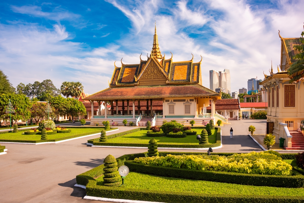 An ornate Cambodian palace with manicured green gardens under a blue sky, with a city skyline behind.